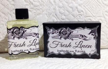 A bottle of Seventh Muse Fragrant Oil in "Fresh Linen" fragrance next to a matching black and white labeled soap bar, both with ornate vintage labels, on a textured white background.