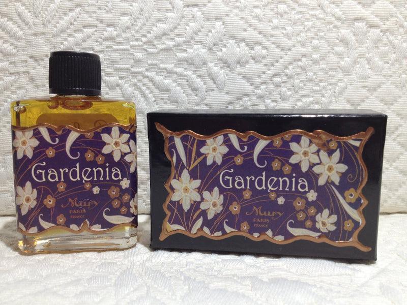 A bottle and a box of "Seventh Muse Fragrant Oil - Gardenia", both featuring intricate floral designs in gold and white on a purple background, set against a textured white fabric.