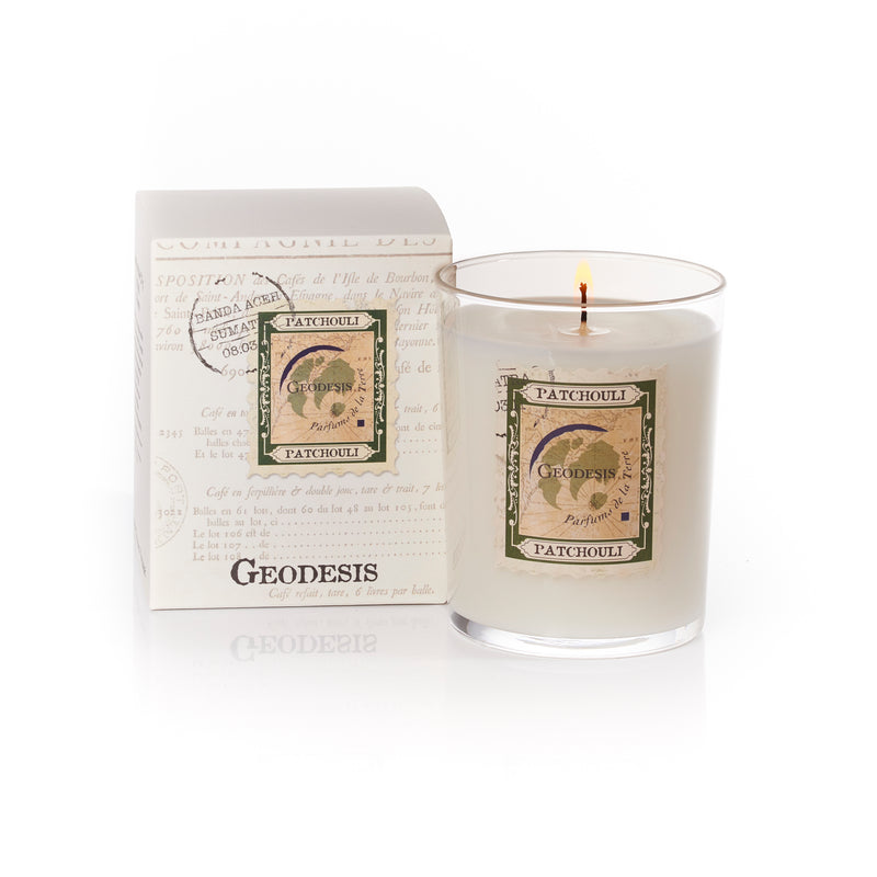 A Geodesis Patchouly 220gm scented candle in a clear glass holder by SCM next to its packaging box with vintage-style labels. The background is pure white, and the woody aroma fills the air.