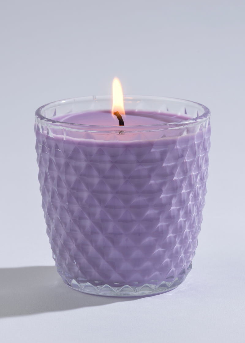 A Sonoma Lavender Soy Wax Round Diamond Glass Candle with a lit wick is inside a glass holder with a textured surface, set against a light gray background.