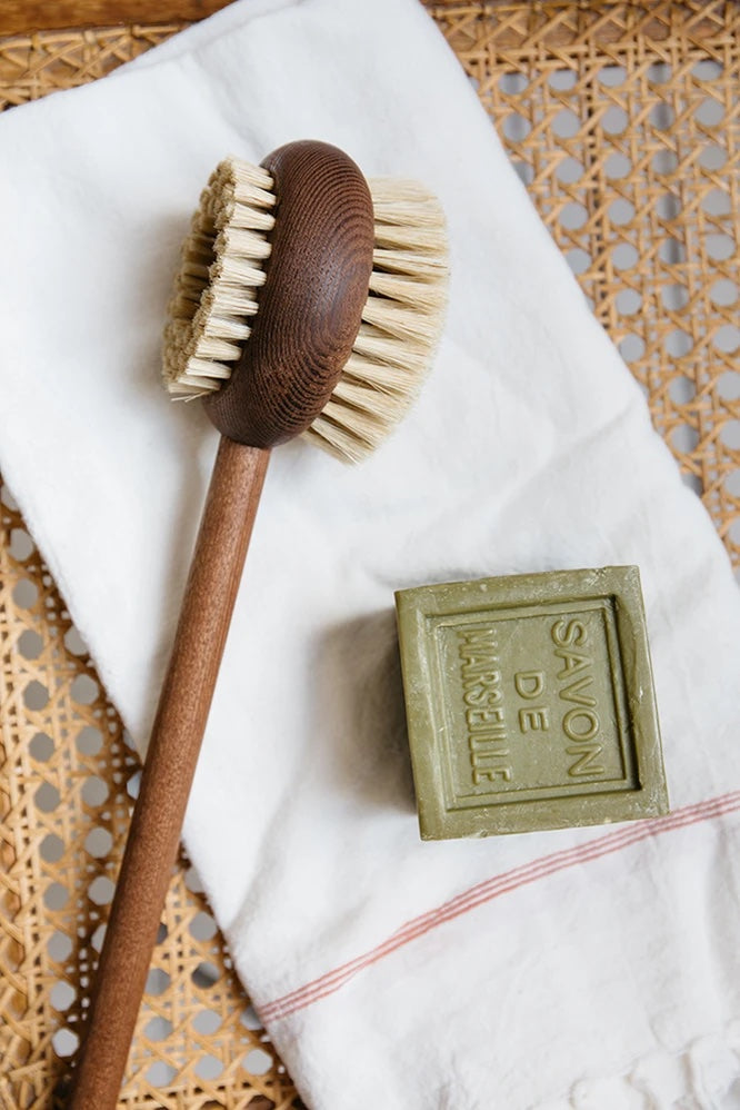 A Andrée Jardin Heritage Ash Wood Handled Body Brush with natural bristles and a bar of green Marseille soap placed on a white towel, which is on a woven rattan surface.