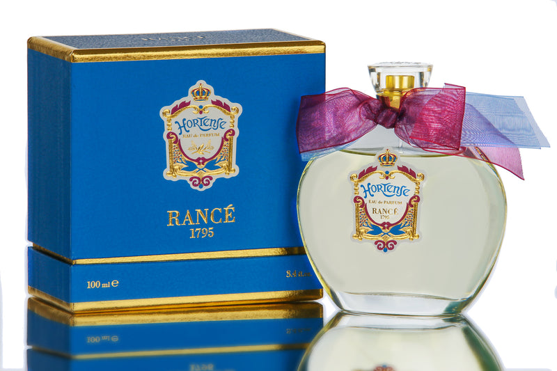 A Rancé Hortense eau de parfum perfume bottle with a purple bow sits next to its elegant blue and gold packaging box on a reflective surface, hinting at the spicy notes it contains.