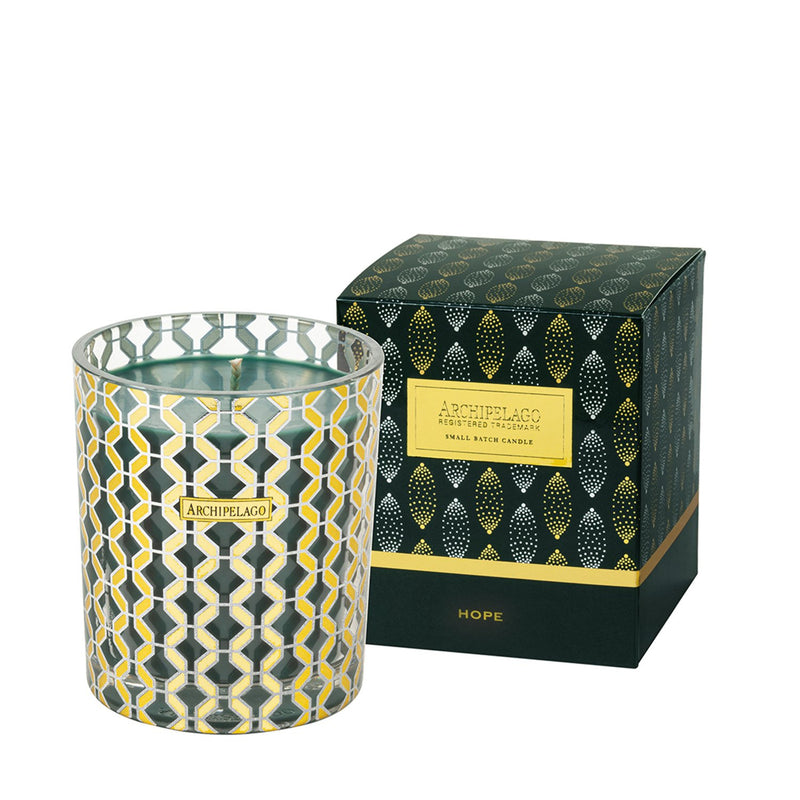 Decorative candle in a glass holder with a geometric pattern beside its elegant black and gold packaging labeled "Archipelago Botanicals," infused with holiday pine.
