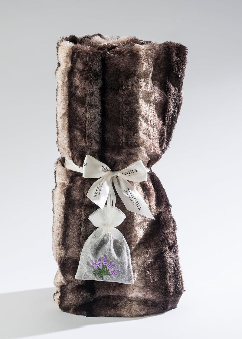 A luxurious Sonoma Lavender Chinchilla Cuddle Throw rolled up and secured with a silky ribbon and a sachet of lavender attached, displayed against a light grey background.