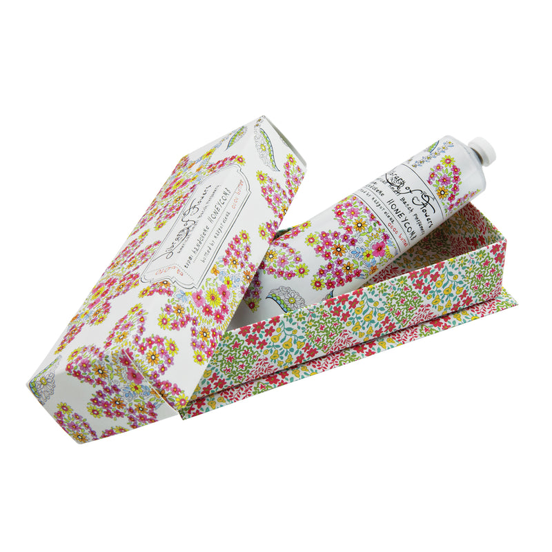 A tube of Library of Flowers Honeycomb Hand Creme, enriched with Shea Butter, partially removed from its Margot Elena floral-patterned packaging, isolated on a white background. The packaging has intricate spring-colored designs with small birds.