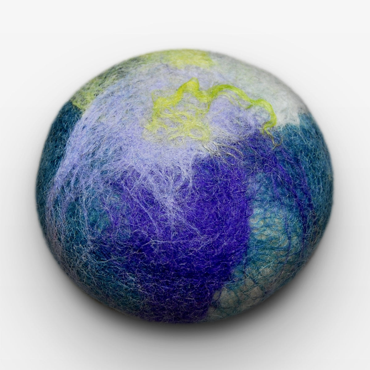 A round, textured object with a surface pattern resembling a Fiat Luxe - Classic Lavender Mint Felted Soap in shades of blue, green, purple, and yellow. The object is centered on a white background.