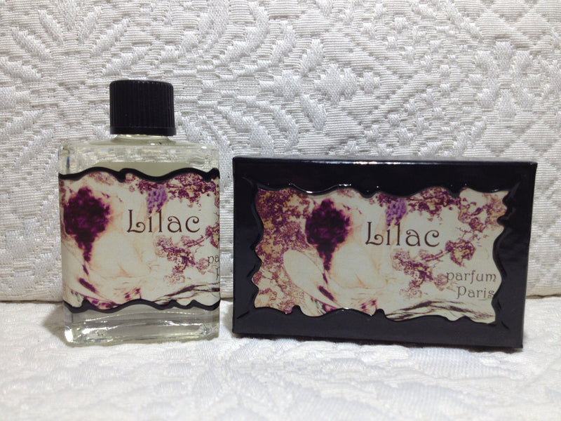 A clear glass perfume bottle labeled "Seventh Muse Fragrant Oil - Lilac" next to its black box with lilac flower designs on a white lace background by Seventh Muse Fragrant Oils.
