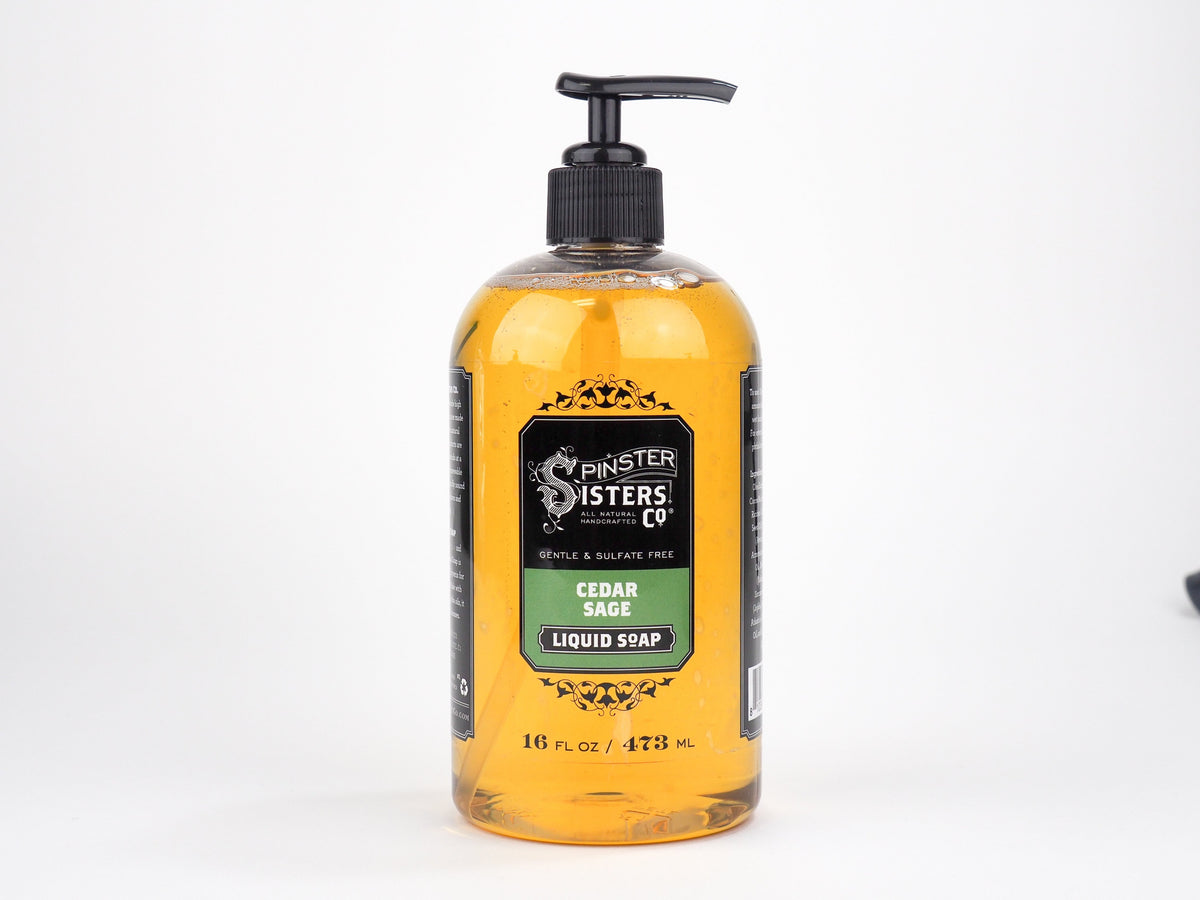 A clear pump bottle of Spinster Sisters, Co. cedar sage liquid soap, enriched with babassu seed oil, filled with yellow liquid, against a white background. The label shows product details.
