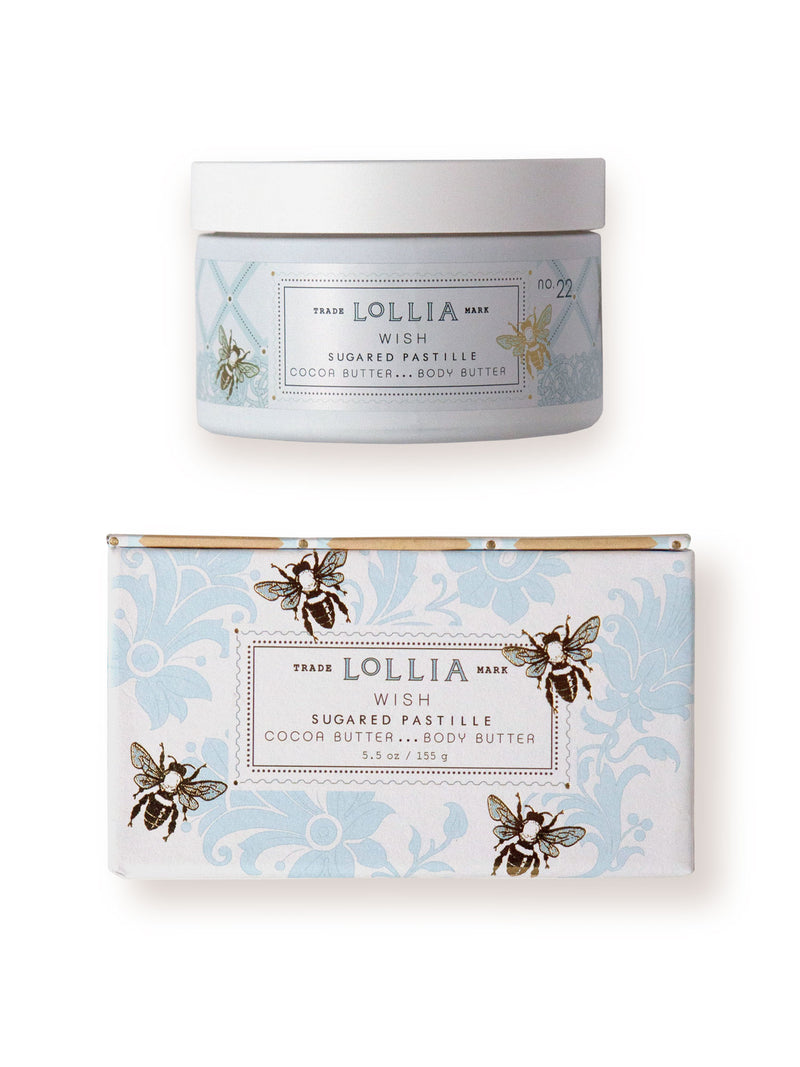 Two Margot Elena Lollia Wish Body Butter products featuring cocoa butter, with elegant light blue packaging adorned with floral designs and bee motifs. One in a tub and the other in a box, displayed against a white