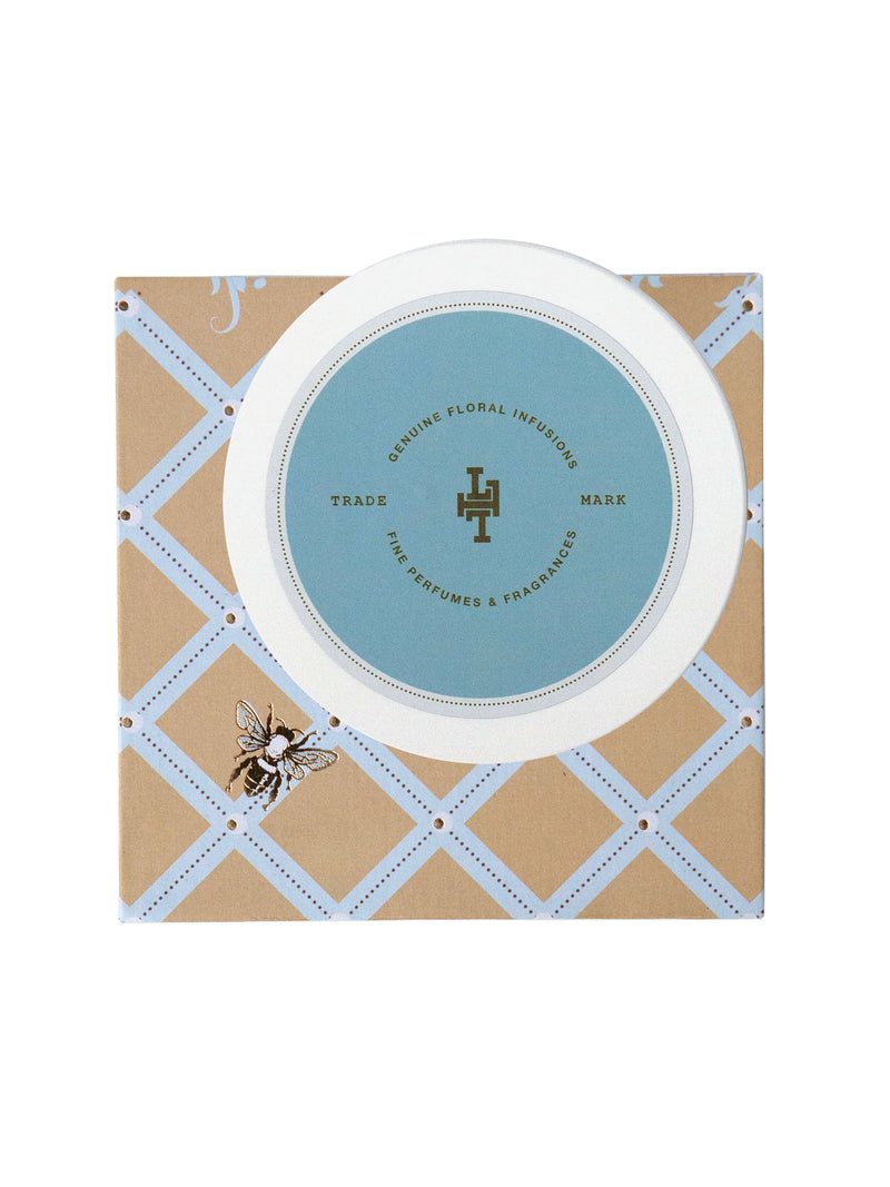 A Lollia Wish Body Butter jar with a light blue lid featuring a Margot Elena logo and text "divine floral infusion" circled by a decorative border, set on a beige and white diagonal stripe patterned background.