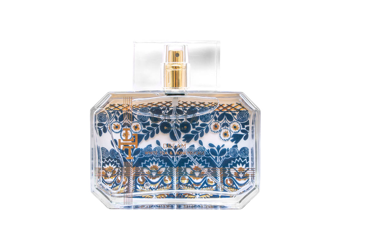 A transparent Lollia Dream Eau de Parfum bottle with intricate blue and gold floral patterns on a white background. The bottle features a rectangular shape and a golden spray nozzle infused with hints of Bergamot, created by Margot Elena.