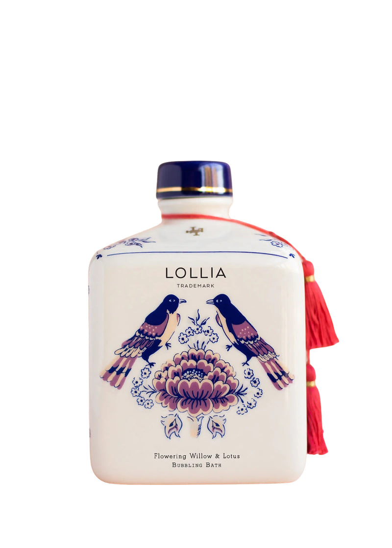 A decorative bottle of Margot Elena's Lollia Imagine Bubble Bath labeled "Flowering Willow & Lotus," featuring blue floral and bird designs with a red tassel hanging from the neck.