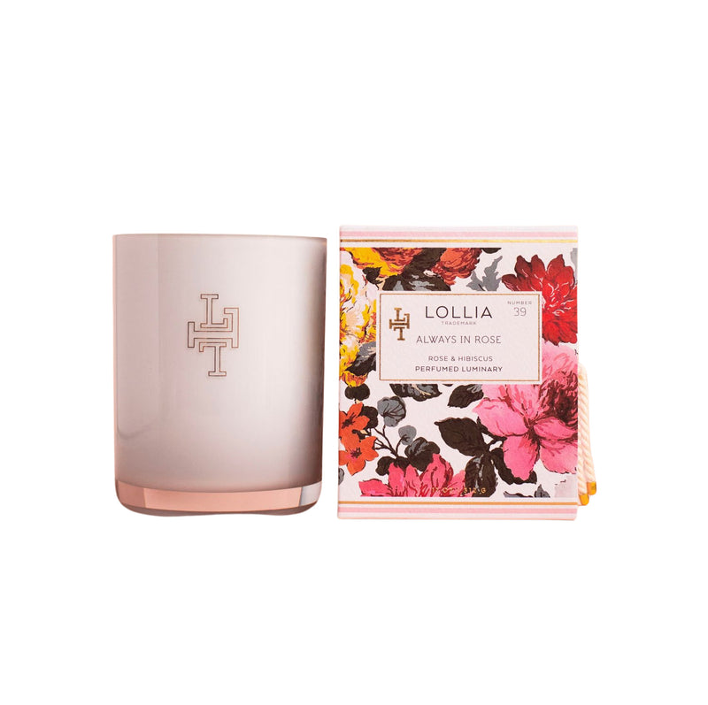 A Lollia Always in Rose No. 39 Perfumed Luminary Candle in a frosted glass container with a modern monogram logo next to its beautifully packaged box adorned with vibrant floral prints in reds and pinks. The box