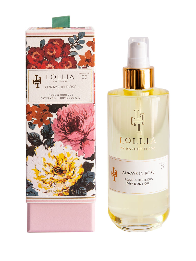 A bottle of Margot Elena's Lollia Always in Rose Dry Body Oil next to its floral-printed packaging box. The box features a dense floral pattern with large pink and yellow blooms.