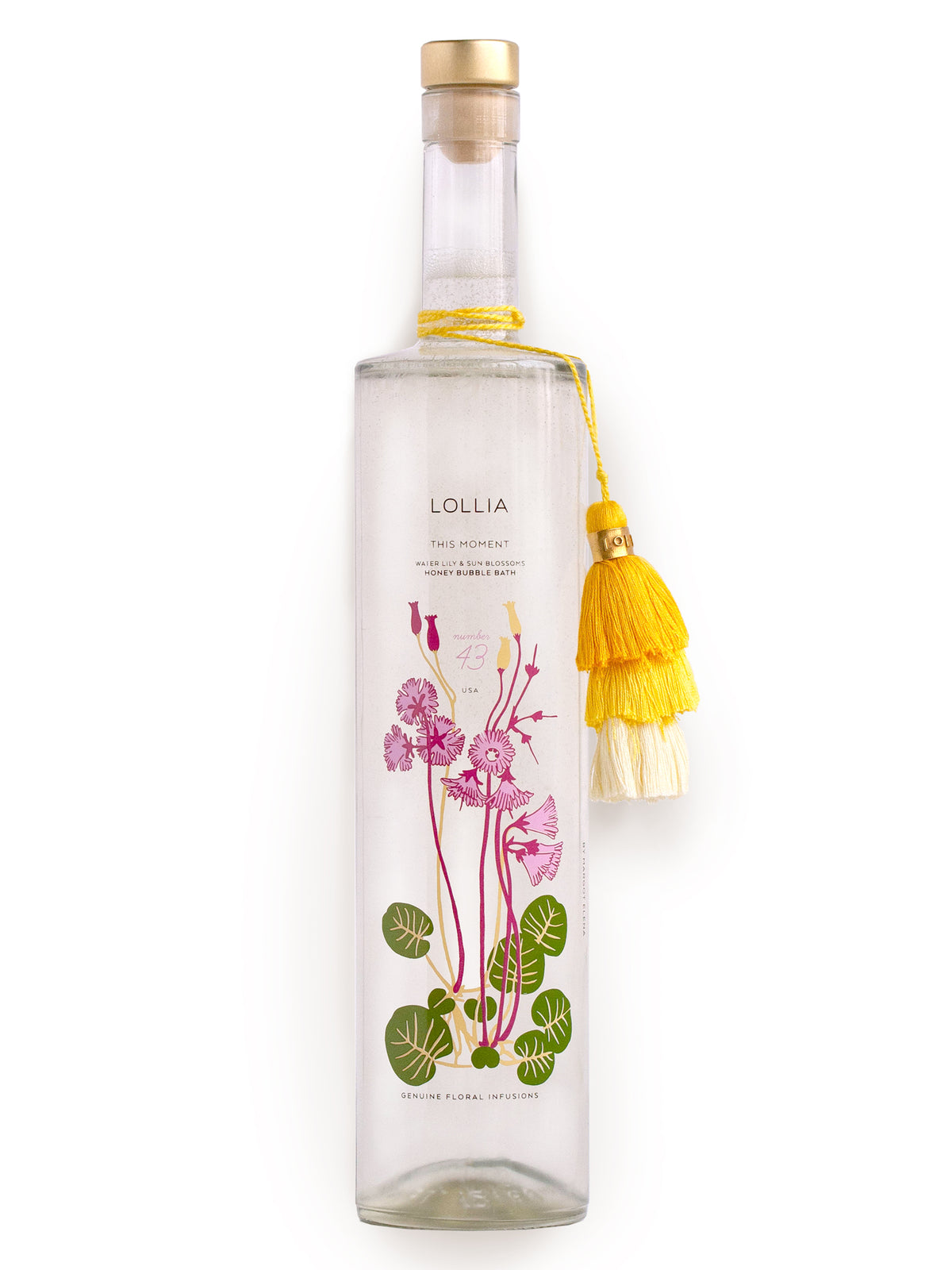 A delicate, clear bottle of Margot Elena's Lollia THIS MOMENT BUBBLE BATH oil with an ornate label featuring pink and green floral illustrations. It's adorned with a yellow tassel on the neck.