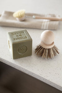 A Marseille soap bar next to an Andrée Jardin Tradition Saucepan Brush with natural bristles, lying on a light grey countertop with a linen towel and spoon in the background.