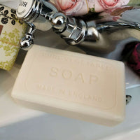 A bar of "The English Soap Co. Vintage Lemon Mandarin Italian Wrapped Soap" made in England resting in a white sink with floral decor in the background.