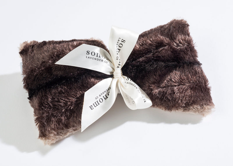 A plush Sonoma Lavender Chinchilla Spa Mask tied with a white ribbon that features elegant, black typography, placed on a bright, clean background infused with aromatic heat.