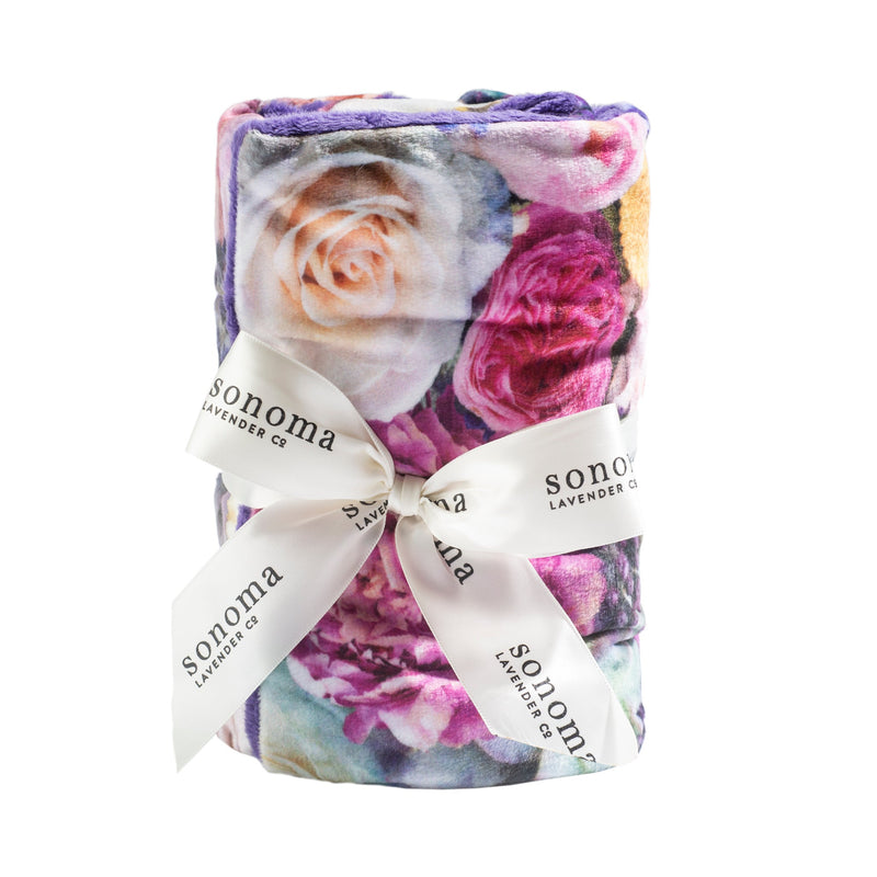 A rolled-up Sonoma Lavender Peony Bouquet Heat Wrap with a floral pattern in shades of purple, pink, and white, tied with a white ribbon printed with "Sonoma Lavender Co." on a white background.