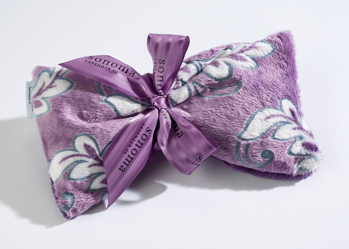 The Sonoma Lavender Violetta Spa Mask features a plush lavender fabric with a soft, fuzzy texture and is adorned with a patterned design of white and green leaves. Infused with aromatic heat to enhance its calming effect, the spa mask is elegantly wrapped with a purple ribbon that displays the brand name "Sonoma Lavender" in black text.