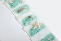 A decorative piece featuring rows of Sonoma OceanAire Sachets by the Yard with a sea salt scent, interspersed with a small shell, a sea snail, a starfish and a seahorse, arranged on.