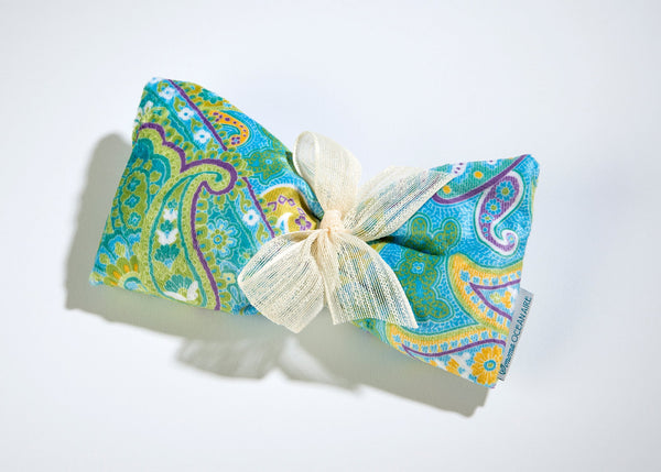 Displayed against a white background, the Sonoma Ocean Aire Palm Beach Paisley Sinus Mask by Sonoma Lavender features a vibrant fabric bundle in blue, green, and yellow paisley patterns. Tied with a cream ribbon, this product is perfect for crafting or creating your own spa mask to relieve tension, bringing comfort and style together.