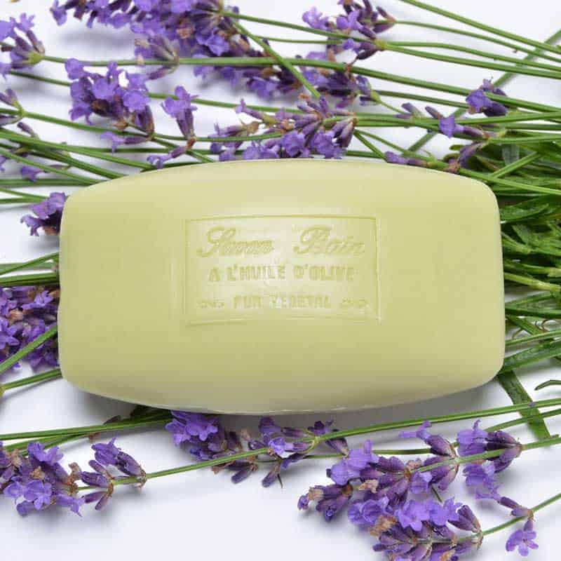 A bar of pale green, moisturizing soap labeled "La Lavande Olive Oil Lavender French Soap" rests on a bed of vibrant lavender flowers with a white background.