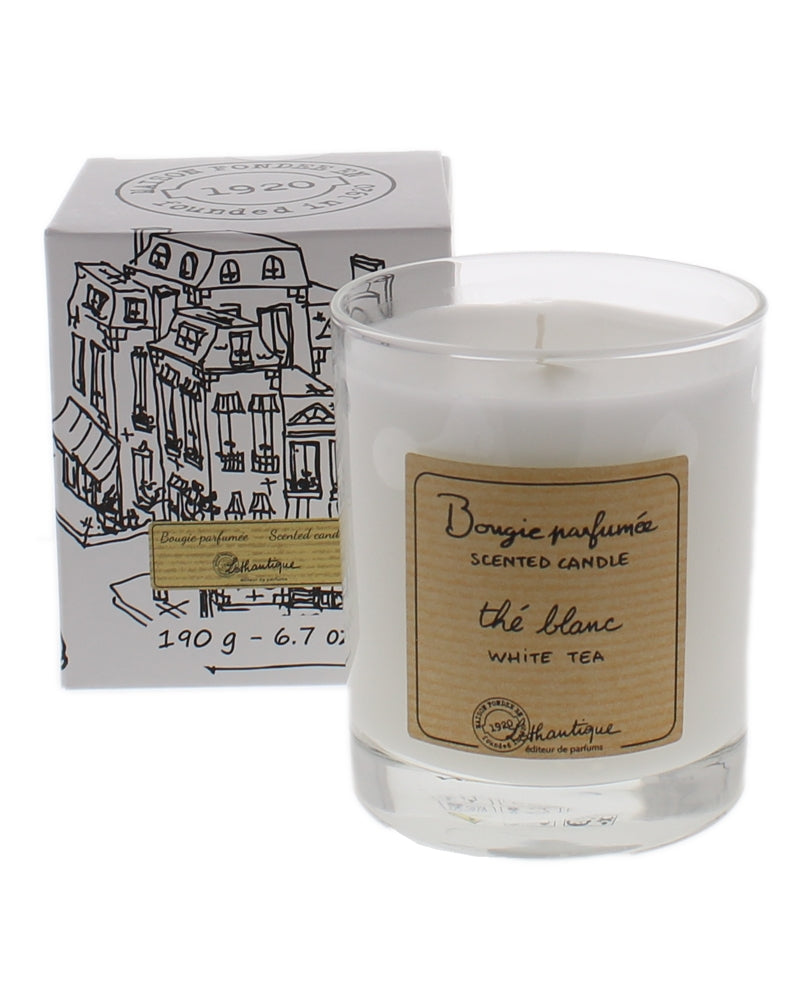 A Lothantique White Tea Candle in a clear glass container labeled "bougie parfumée, thé blanc" beside its illustrated box, depicting urban storefronts.