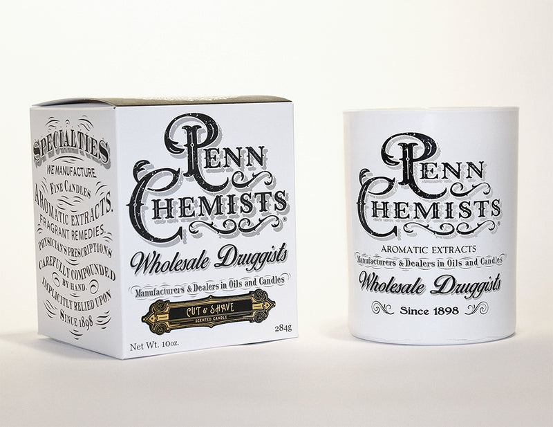 Two vintage-style barber shop containers: one a square box and the other a cylindrical bottle, both with ornate black and white labels featuring text about Penn Chemists Classic Candle - Cut & Shave and establishment in 1898.