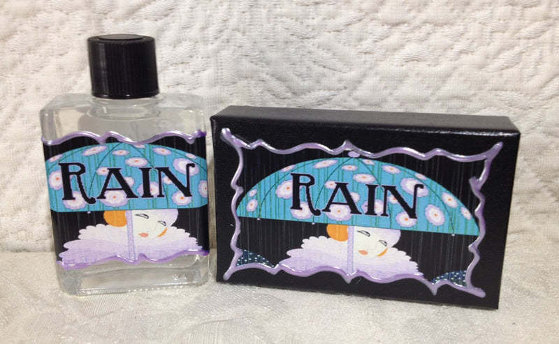 A clear perfume bottle and a black box, both decorated with illustrated labels featuring a whimsical design of a person under a purple umbrella with "rain" written above. Background of textured fabric is enhanced by Seventh Muse Fragrant Oil - Rain from Seventh Muse Fragrant Oils.