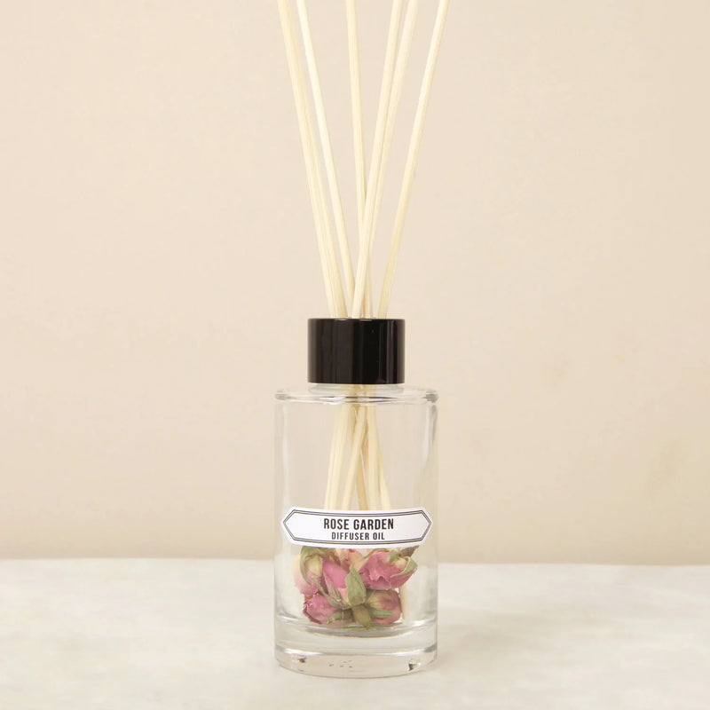 A clear glass diffuser bottle labeled "Norfolk Natural Living Rose Garden diffuser" with pink rosebuds inside and several reed sticks inserted, set against a neutral background.