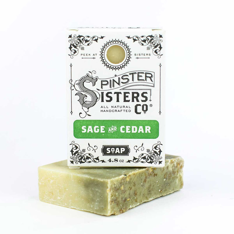 A bar of Spinster Sisters Co. handmade, all-natural vegan Sage & Cedar with Oatmeal bath soap, with visible botanical inclusions, displayed in front of its packaging on a white background.