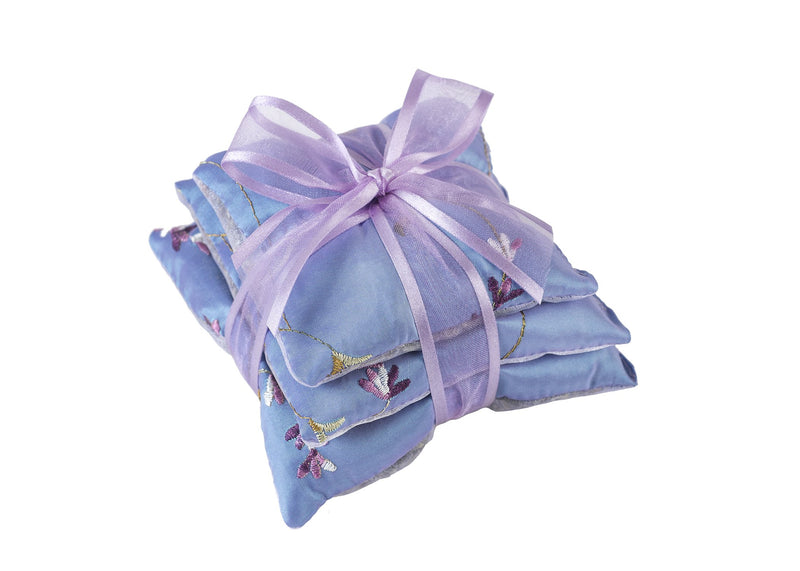 A decorative light blue pillow with a subtle floral pattern, adorned with Sonoma Lavender Sachet Trios - Embroidered Lavender and a translucent lilac ribbon tied in a graceful bow on top, isolated against a white background.