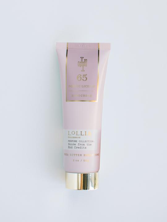 A tube of Lollia Poetic License Score From the End Credits Hand Creme with hydrating shea butter, featuring a pink and gold design, lying against a plain white background by Margot Elena.