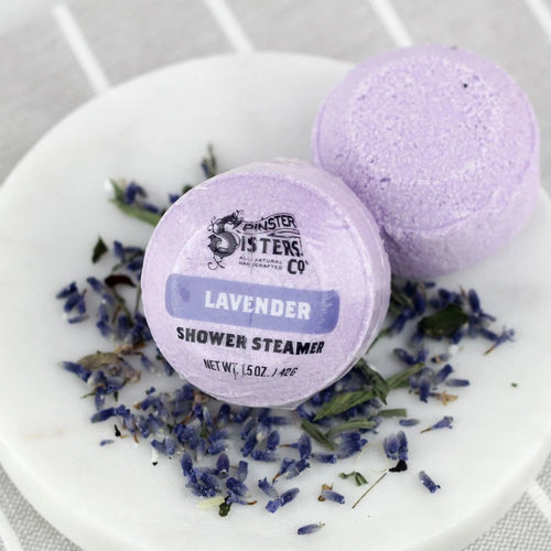 Two Spinster Sisters Lavender Shower Steamers on a white plate with dried lavender buds scattered around, one is labeled "lavender essential oil shower steamer.