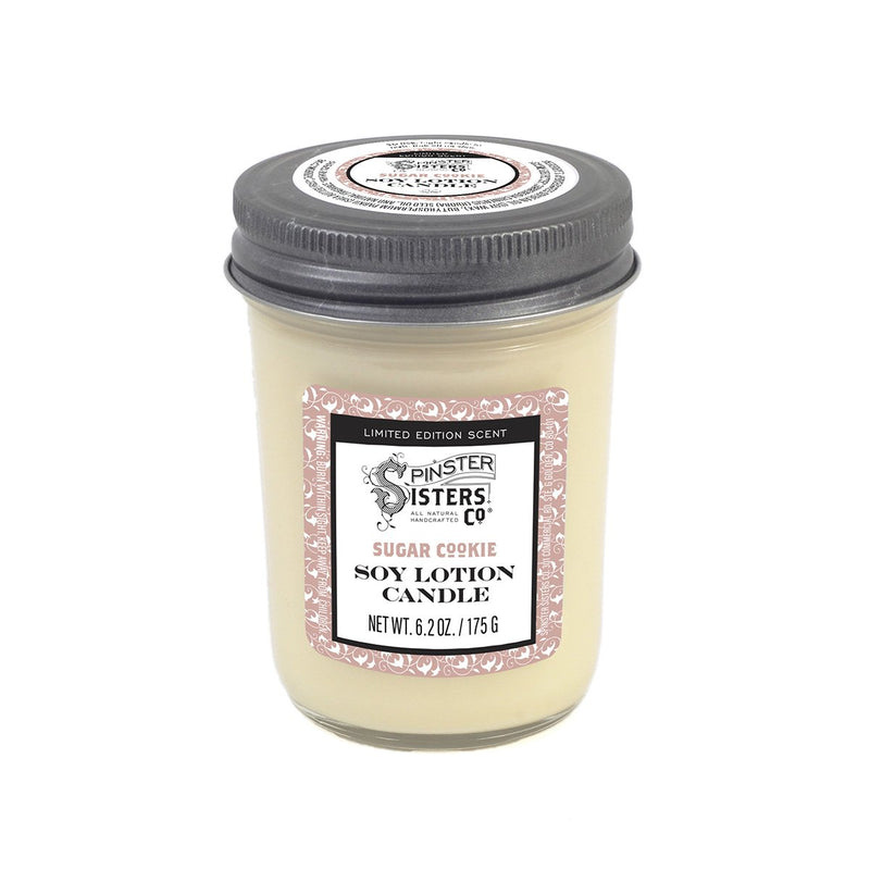 A glass jar of Spinster Sisters, Co. soy lotion candles labeled "Spinster Sisters Soy Candle - Sugar Cookie", with a cream color and a white lid, set against a plain background.