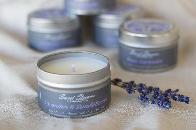 A lit Sweet Streams Lavender Co. - Pure Lavender Sandalwood Soy Travel Tin Candle sits in the foreground. Several unlit candles and sprigs of lavender are blurred in the background on a soft fabric surface.