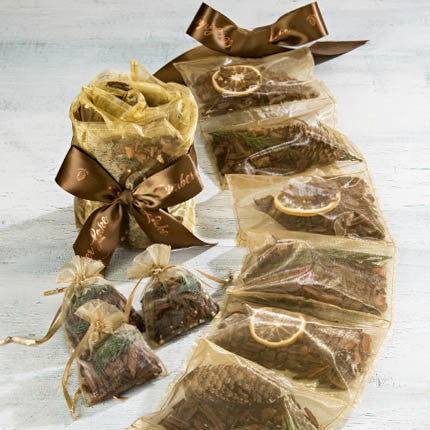 Six transparent gift bags arranged in a line, each filled with natural Sonoma Timber Lake sachets by-the-yard ingredients such as dried flowers and citrus slices, tied with brown ribbons.