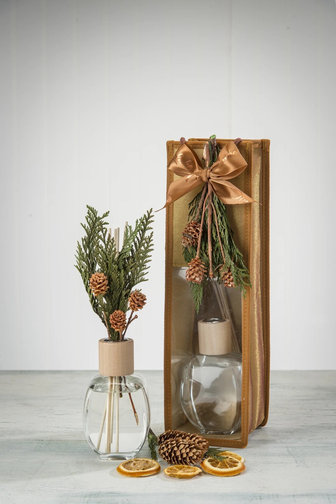 A holiday-themed still life featuring a glass vase with evergreen branches and pine cones, a Sonoma Timber Lake Diffuser from Sonoma Lavender, and dried orange slices on a wooden surface.