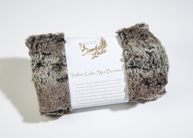 A pair of fluffy gray Sonoma Lavender heated spa booties with a label reading "Sonoma Timber Lake Woodland Fur Spa Booties" laid on a light background.