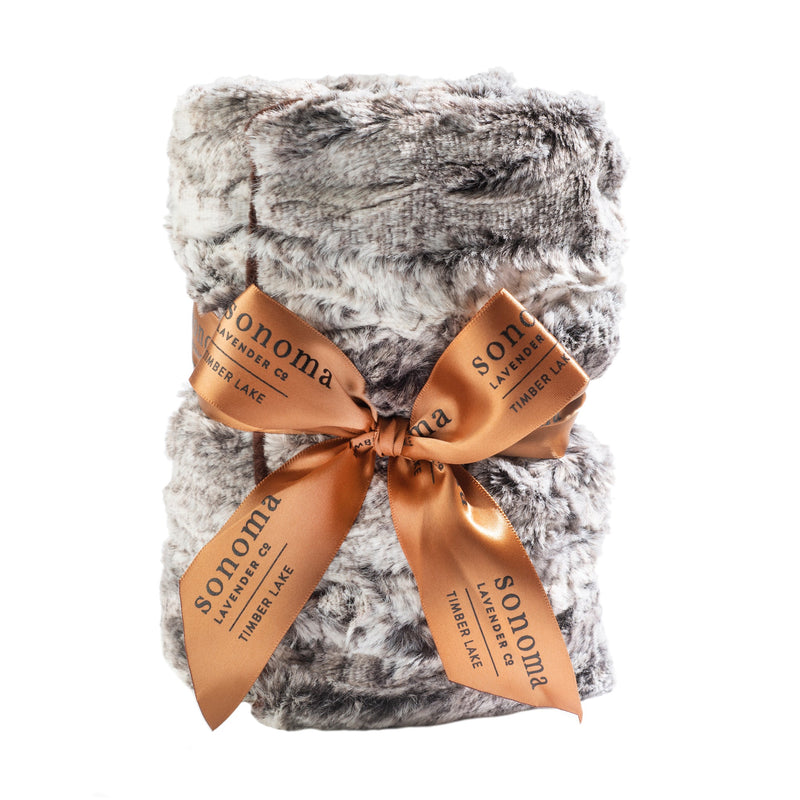 A plush, gray faux fur blanket neatly folded and tied with a shiny, copper-colored ribbon featuring the word "Sonoma Lavender" printed in white. The background is pure white, hinting at the comforting Sonoma Timber Lake Winter Frost Heat Wrap.