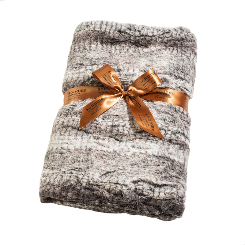 A plush gray and white Sonoma Lavender blanket neatly folded and tied with a shiny copper ribbon, infused with the aromatic, mood-elevating scent of Timber Lake Winter Frost, labeled "Sonoma" on an isolated white background.