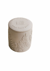 A cylindrical, textured ceramic container with a lid, featuring embossed designs of plants and the phrase "static beauty world" on the top. The background is solid white, and it holds a Margot Elena Himalayan Salt & Rosewood Ceramic Candle.