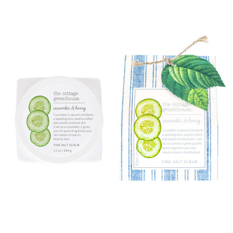 Two packages of The Cottage Greenhouse Cucumber & Honey Fine Salt Scrub with avocado oil displayed on a blue striped cloth, one open showing the product with cucumber slices and leaves by Margot Elena.