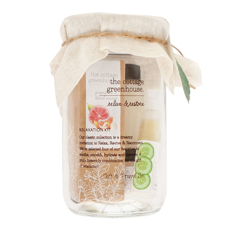 A clear glass jar with a beige fabric lid cover, labeled "Margot Elena Cottage Greenhouse Fruit Relaxation Kit," displaying cucumber and mint products inside, including a facial mask and bubbling milk bath.