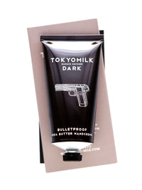 A tube of Margot Elena TokyoMilk Dark Bulletproof Handcreme, featuring a graphic of a pistol, positioned against its own packaging box.