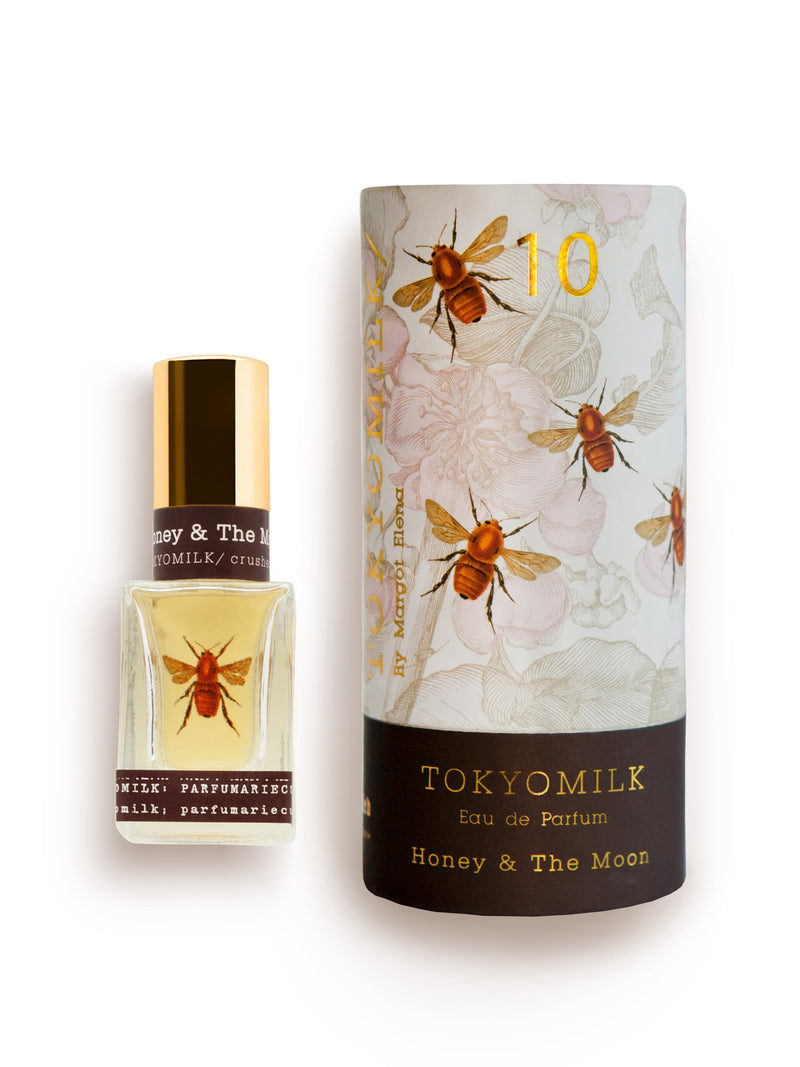 A bottle of Margot Elena TokyoMilk Honey & The Moon No. 10 Parfum next to its packaging, both adorned with artistic floral and bee designs on a white background, featuring