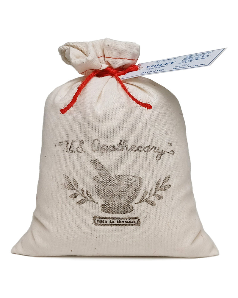 A beige cloth gift bag labeled "U.S. Apothecary" with a red drawstring and a blue tag, featuring an image of a mortar and pestle and the text "est. U.S. Apothecary Violet + Yarrow Bath Soak Bag".