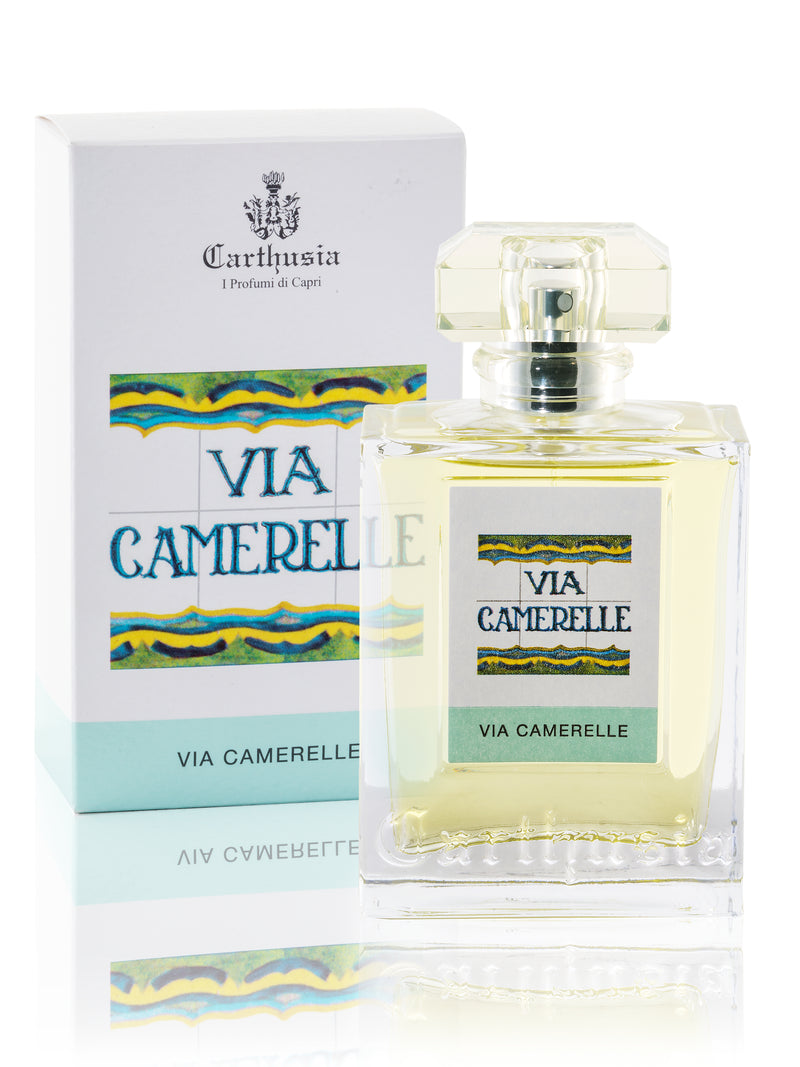 A glass perfume bottle labeled "Carthusia Via Camerelle Eau de Parfum - 100ml" in front of its matching packaging box. The perfume is clear, and the design features wavy blue and green lines on a white background influenced by Carthusia I Profumi de Capri.