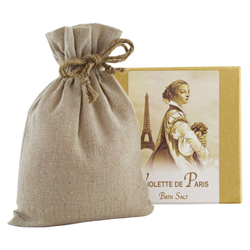 A canvas pouch tied with a jute string next to a box of La Bouquetiere Violette de Paris Bath Salts with Linen Bag, featuring a vintage illustration of a woman holding flowers with the Eiffel Tower in the.